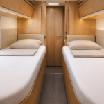 Camera posteriore di un camper con due letti gemelli, ciascuno con lenzuolo con angoli bianco in cotone ben teso e un cuscino, tra mobili in legno chiaro e un finestrino
